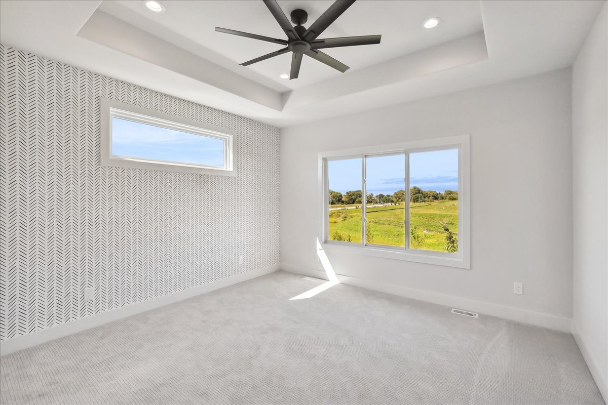 A bright, empty room with a white and gray herringbone accent wall, large windows showing a green landscape, a tray ceiling with a black ceiling fan, and light gray carpeting.