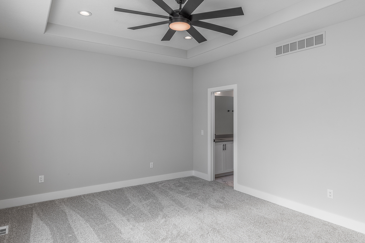 A modern empty room featuring light gray walls, carpeted floors, and a large ceiling fan with multiple blades. There is a white door leading to an adjacent space with a glimpse of a sink and countertop. Recessed ceiling lights add to the contemporary feel.