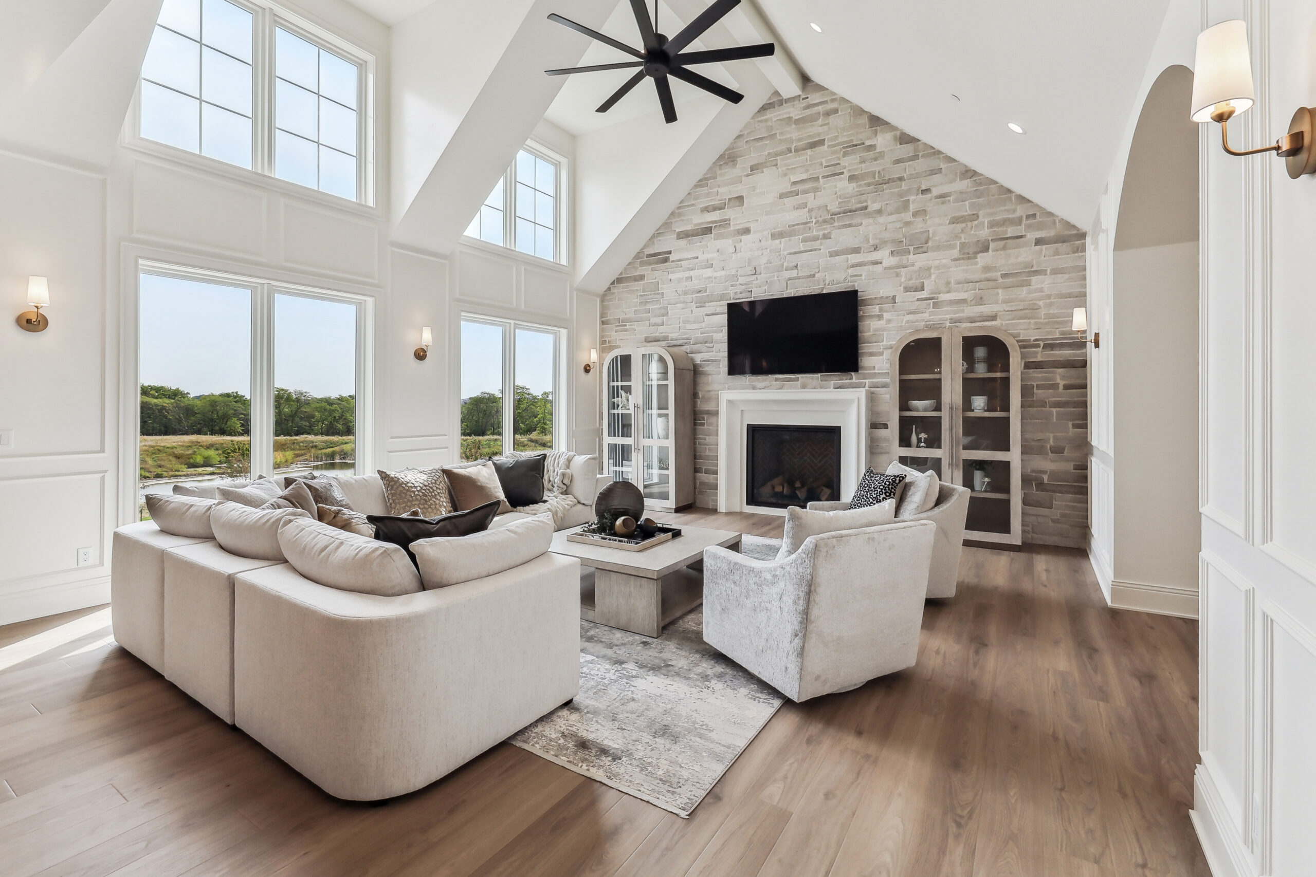 A spacious living room featuring a large L-shaped sectional, two armchairs, and a coffee table. The room has high ceilings, a ceiling fan, and a stone fireplace with a mounted TV. Large windows offer a view of greenery outside.