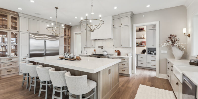 A modern kitchen with a large island, surrounded by four cushioned chairs. The kitchen features stainless steel appliances, white cabinets, wooden flooring, and elegant pendant lighting. A pantry with open shelving is visible in the background.