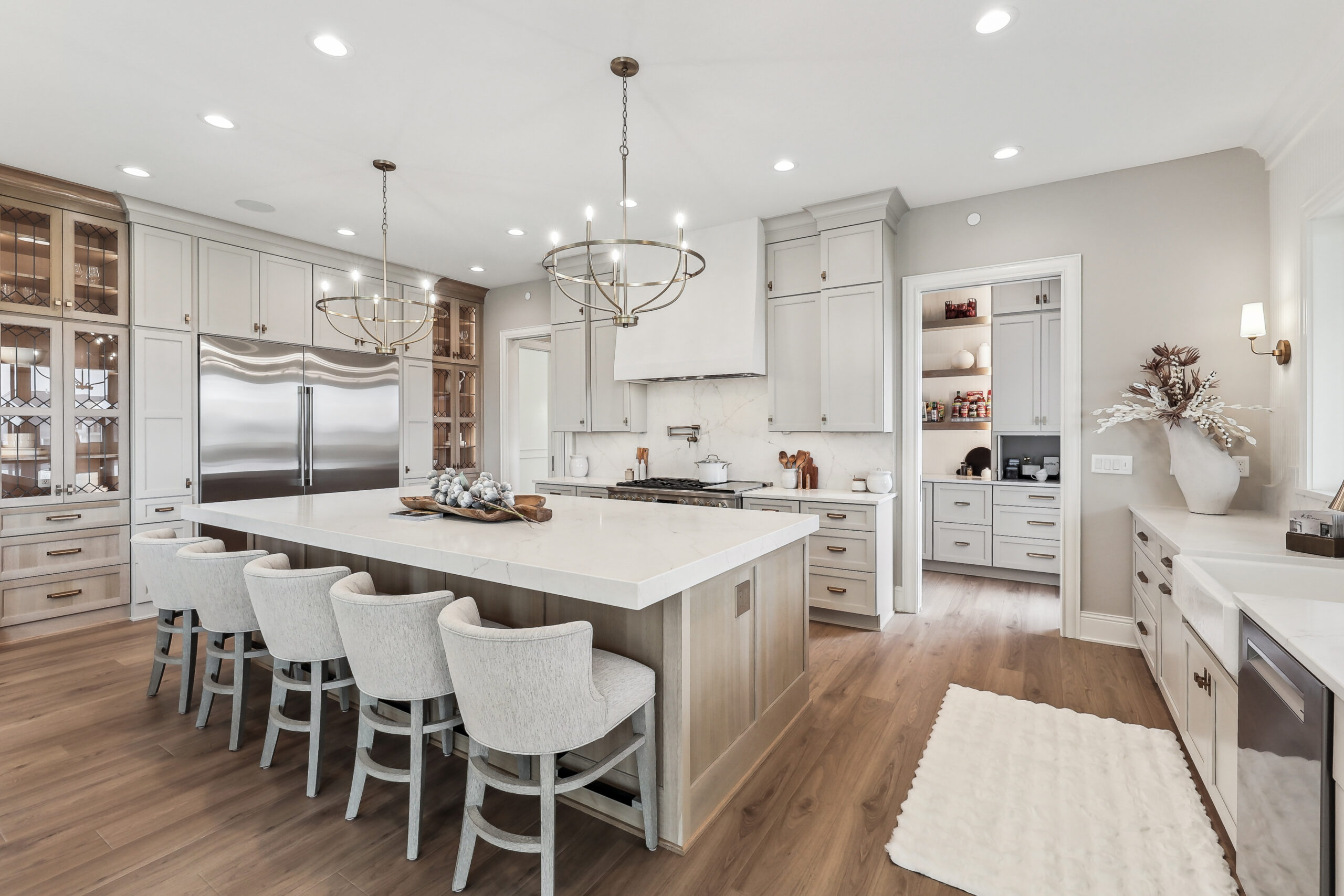 A spacious kitchen featuring a large island with white countertops and four cushioned stools. Modern chandeliers hang above the island. Stainless steel appliances, light wood floors, and white cabinetry complete the elegant look.