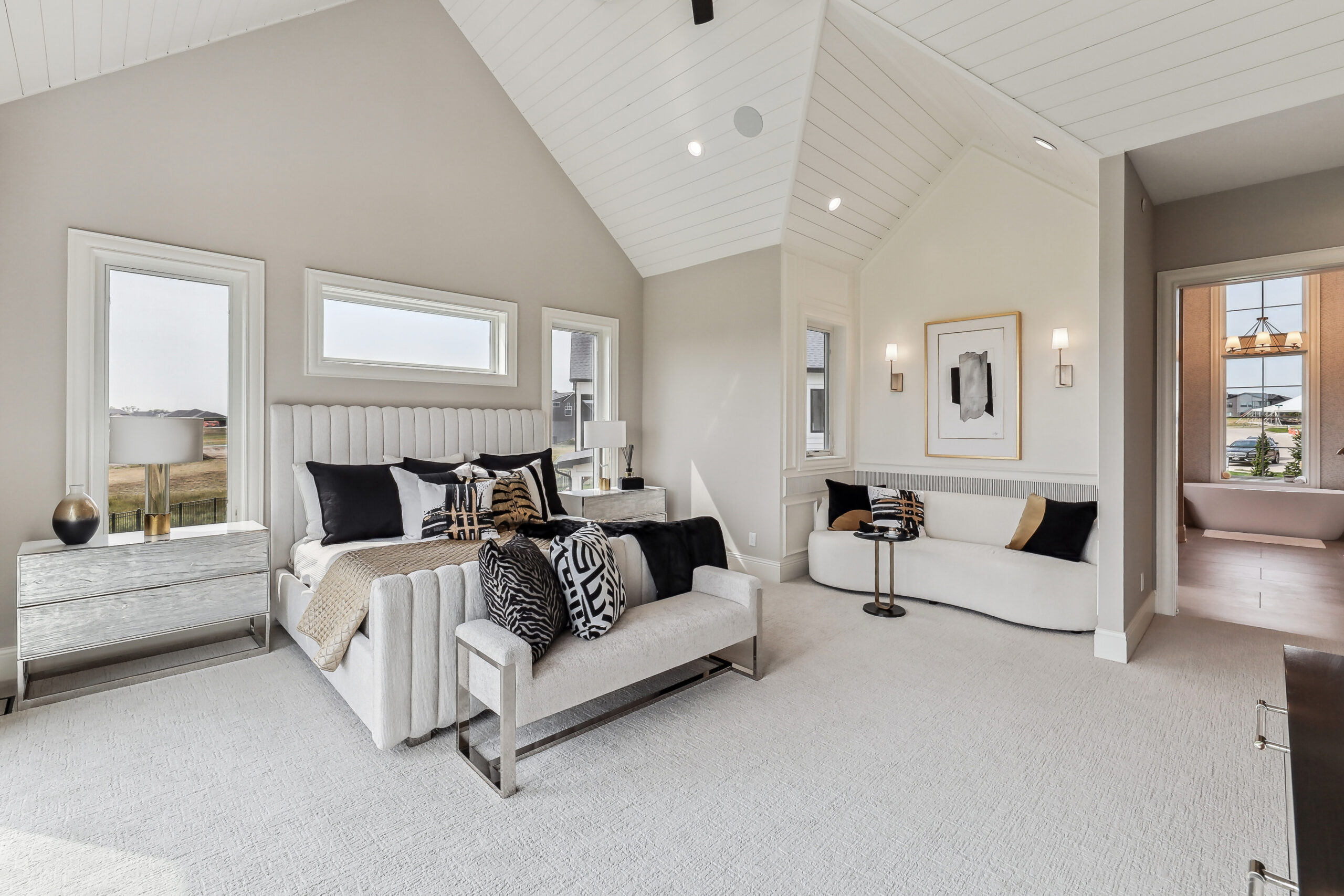 A spacious bedroom with vaulted white shiplap ceilings, light carpeting, and three large windows. The room features a double bed with white and black bedding, a white bench at the foot, a small sitting area with a couch and table, and modern wall art.