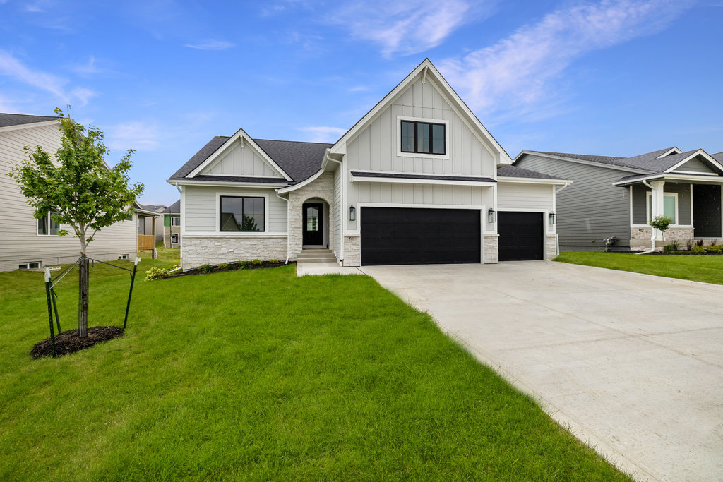 A modern, single-story house with light gray siding and a dark roof. It features a large, three-car garage with dark doors, a well-manicured lawn, and a young tree. The sky is clear and blue.