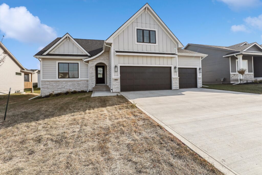 A modern single-story house with a light beige exterior and stone accents, featuring a dark double garage door. It sits on a neatly maintained lawn with a wide driveway, under a clear blue sky.