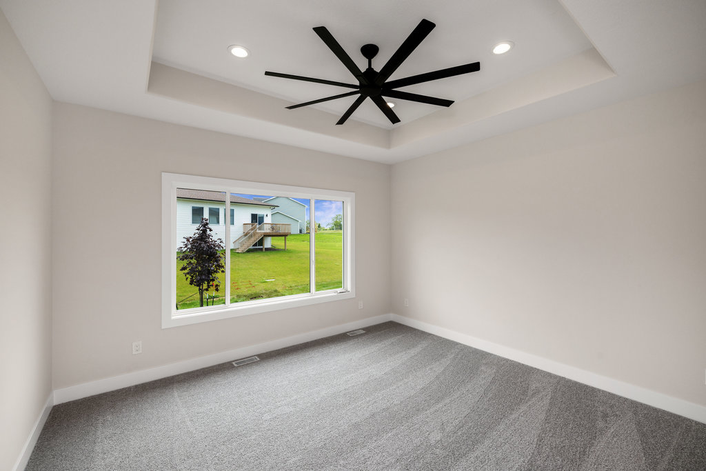 An empty room with light gray carpet and white walls. A large window reveals a view of a green yard and a house. A modern black ceiling fan is centered in the room, and recessed lighting is installed in the ceiling.