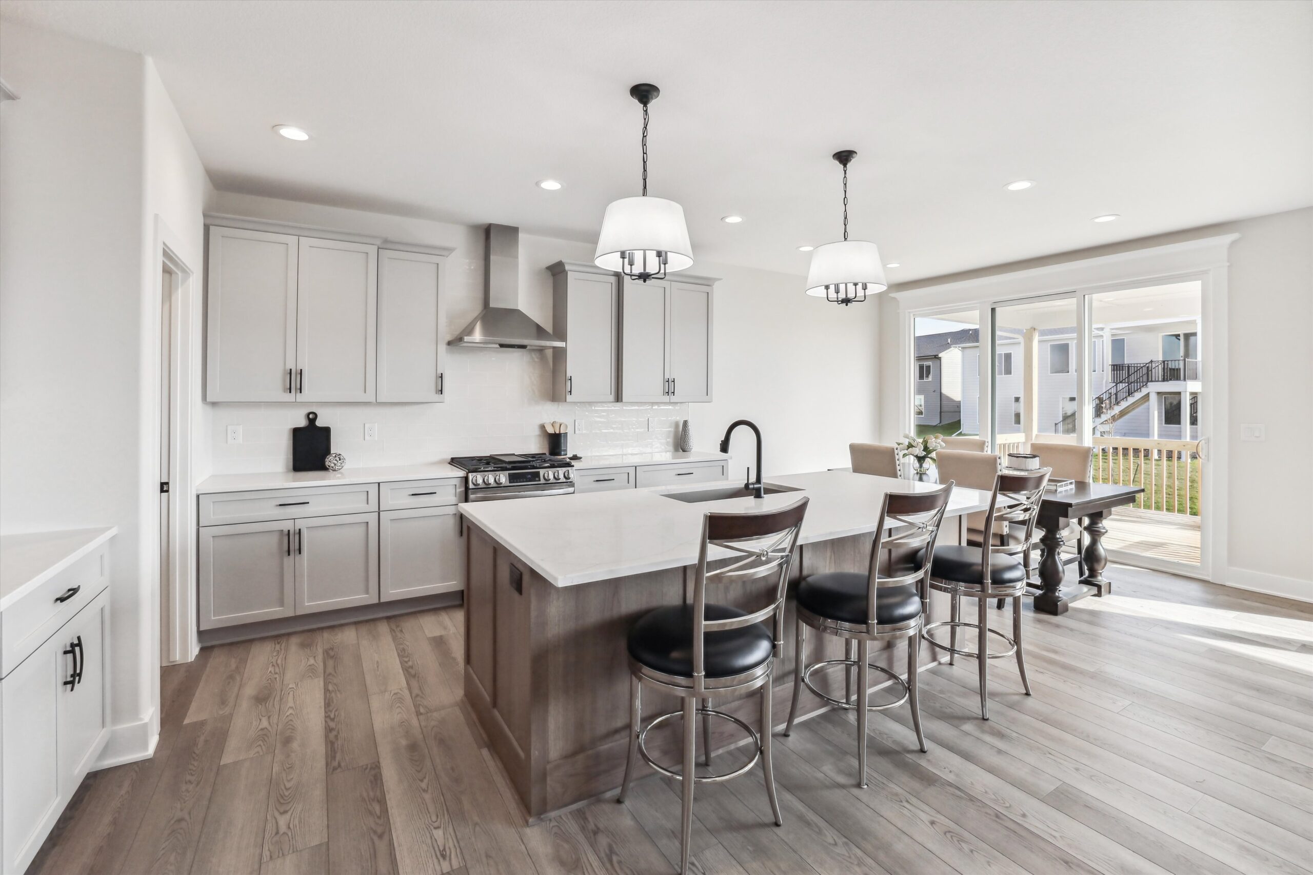 A modern kitchen with light gray cabinets and a central island with bar stools. Stainless steel appliances, including a stove and a range hood, are featured. Sliding glass doors in the background lead to an outdoor patio, with ample natural light.