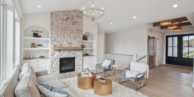 A modern living room with a stone fireplace, light-colored walls, and wooden floors. It features a gray sofa, two gold and white chairs, round gold tables, and a fluffy white rug. A chandelier hangs above, and large windows provide natural light.