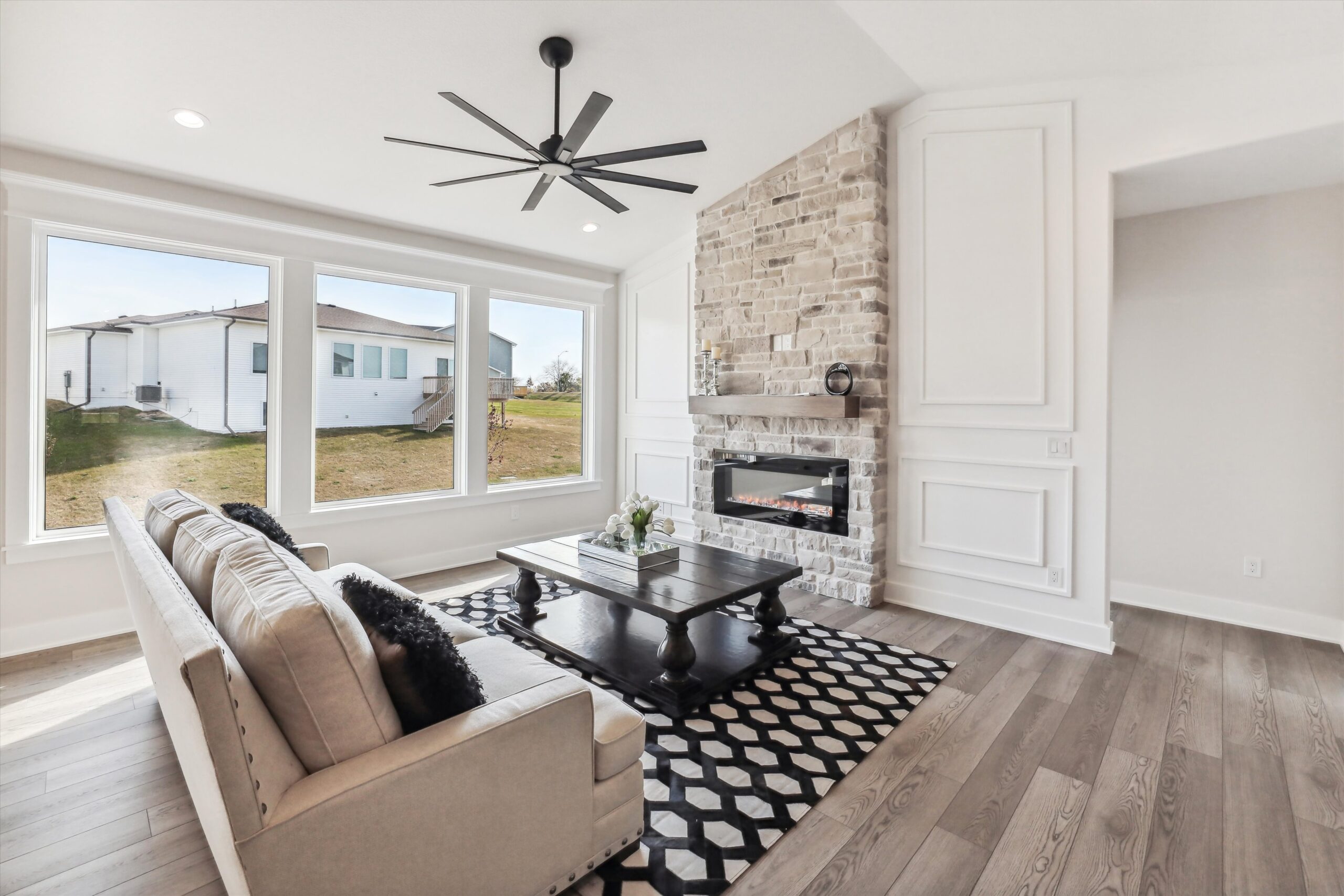 Spacious living room with a beige couch, black and white rug, and a dark wood coffee table. Large windows provide natural light. A stone fireplace with a modern electric insert and a black ceiling fan complete the contemporary design.