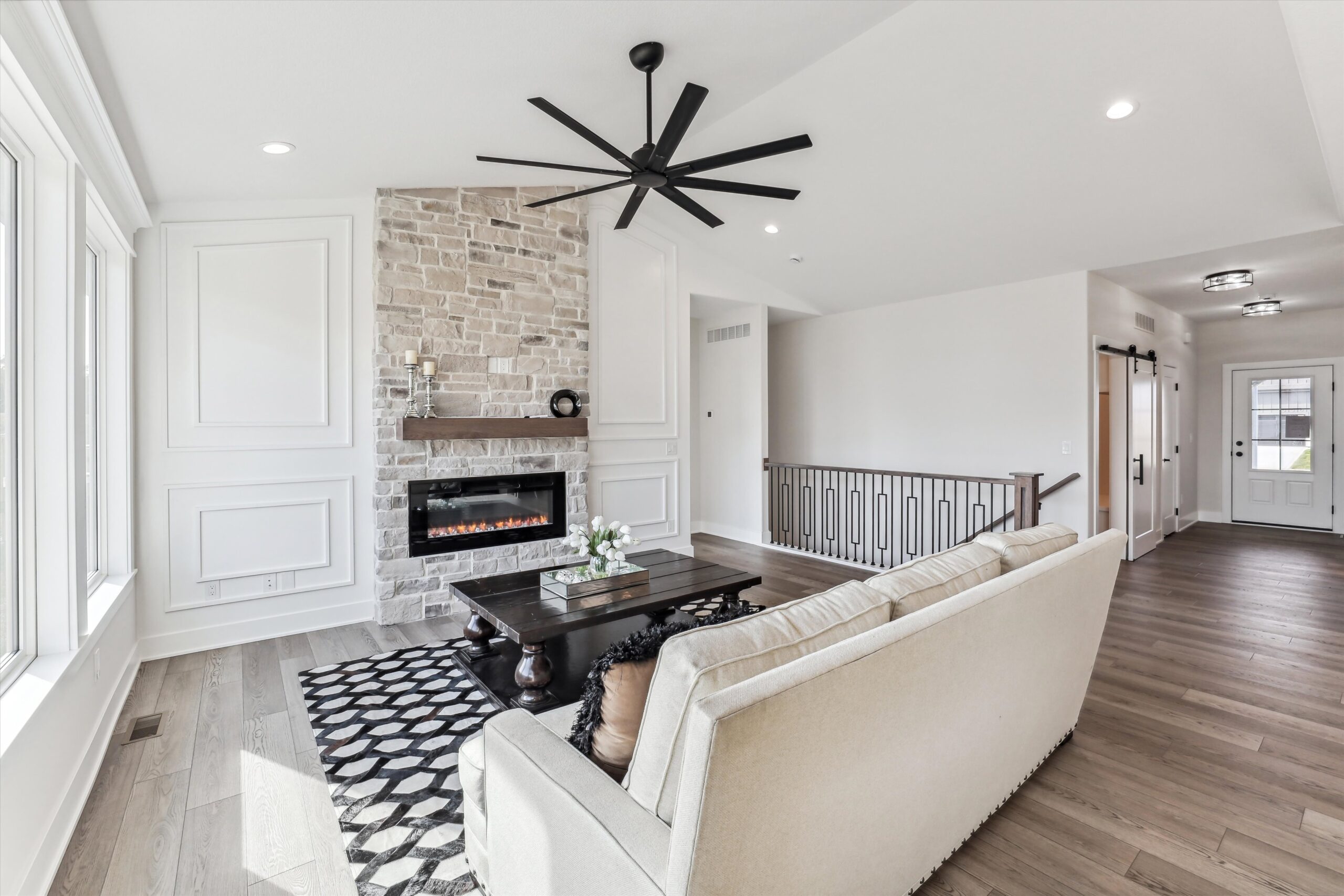 A modern living room features a stone fireplace with a black mantel, a beige sofa, and a black coffee table on a patterned rug. A ceiling fan is above, large windows provide ample light, and a railing leads to a hallway with a white door.