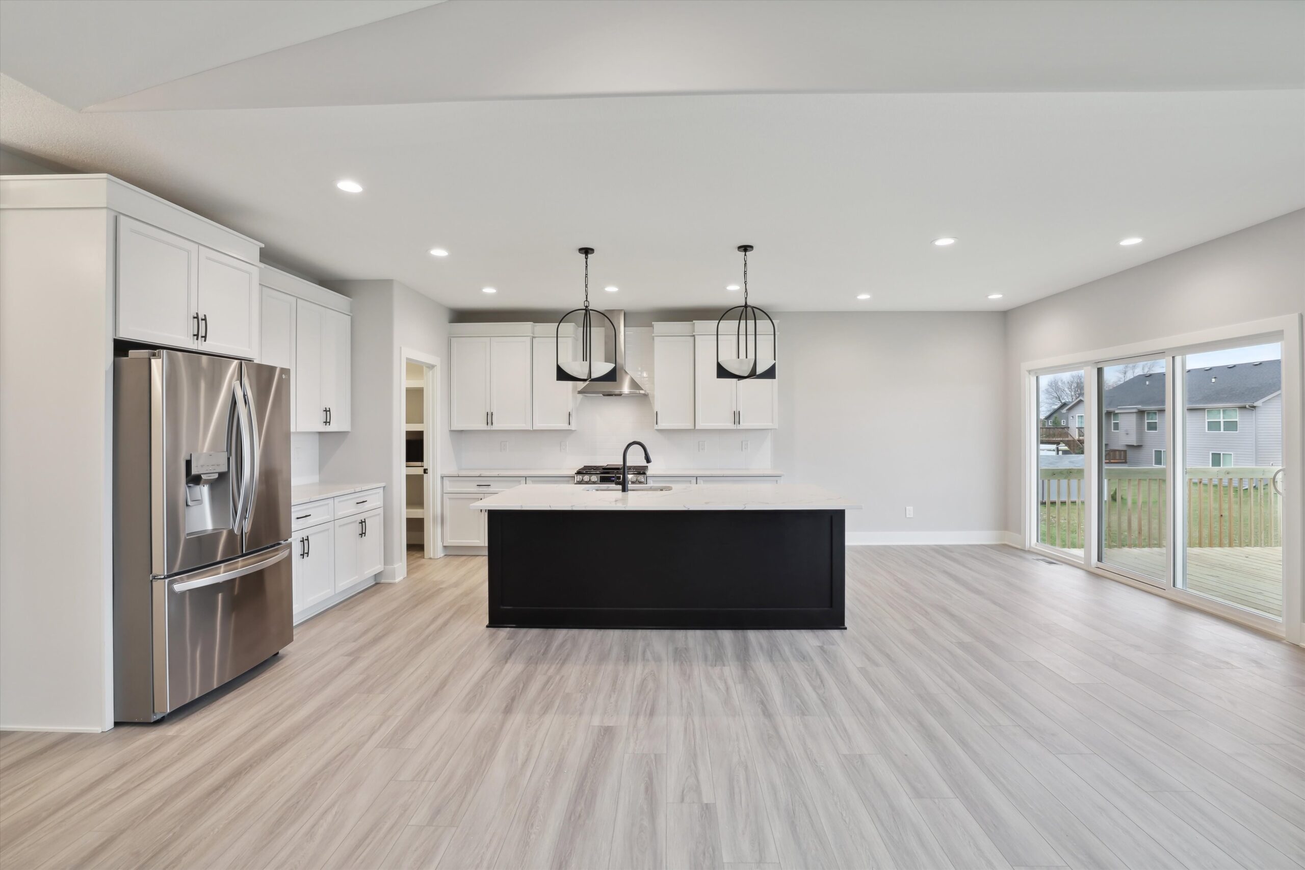 Modern kitchen with white cabinets, stainless steel appliances, and a large black island. Two pendant lights hang above the island. Spacious room with light wood flooring and large sliding glass doors opening to a deck with a view of the backyard.