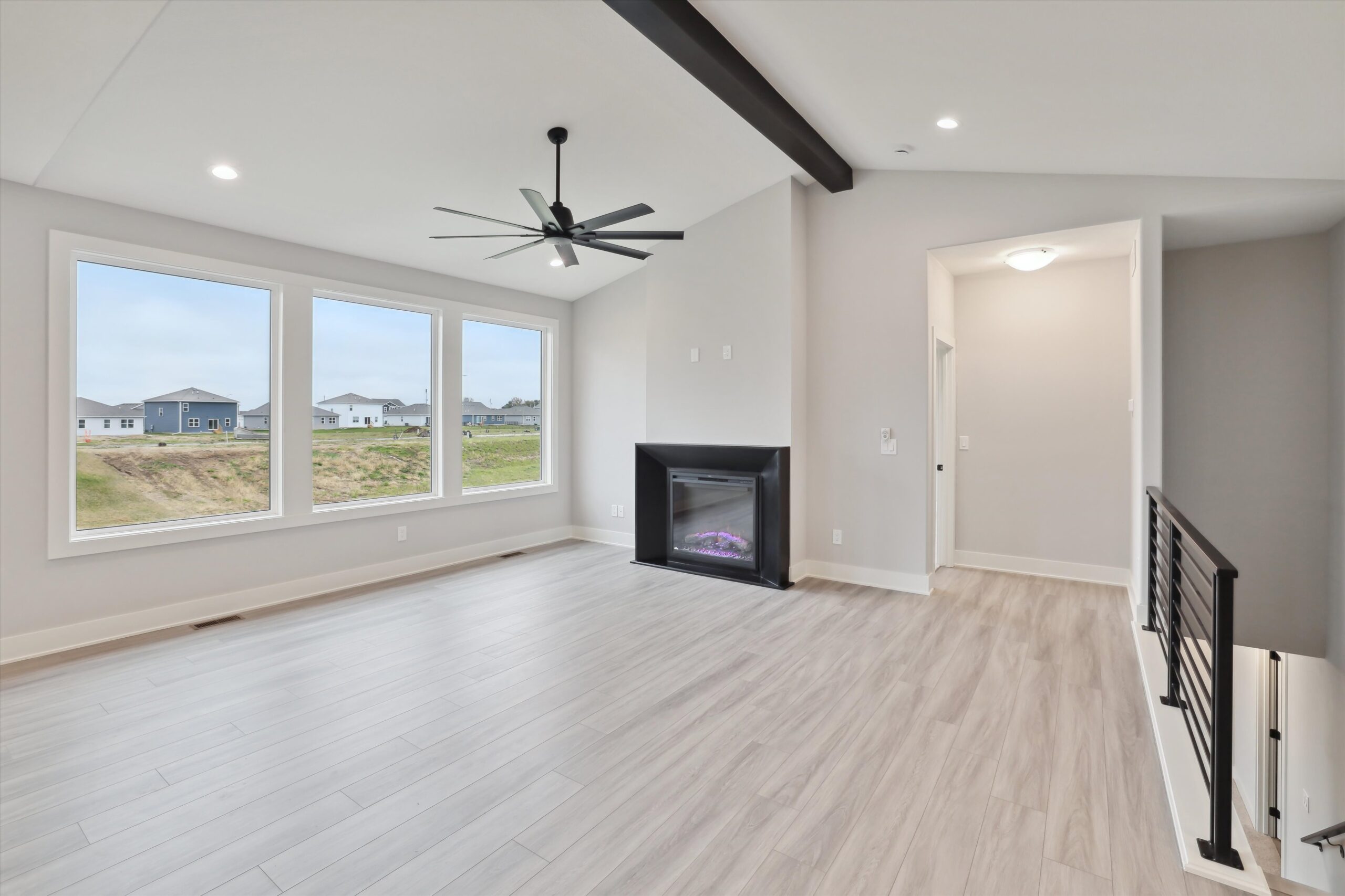 A modern, unfurnished living room with large windows, light wood flooring, and a ceiling fan. A fireplace is centered on one wall, and a staircase with a black railing is visible to the right. Outside, houses and grassy lawns are seen through the windows.