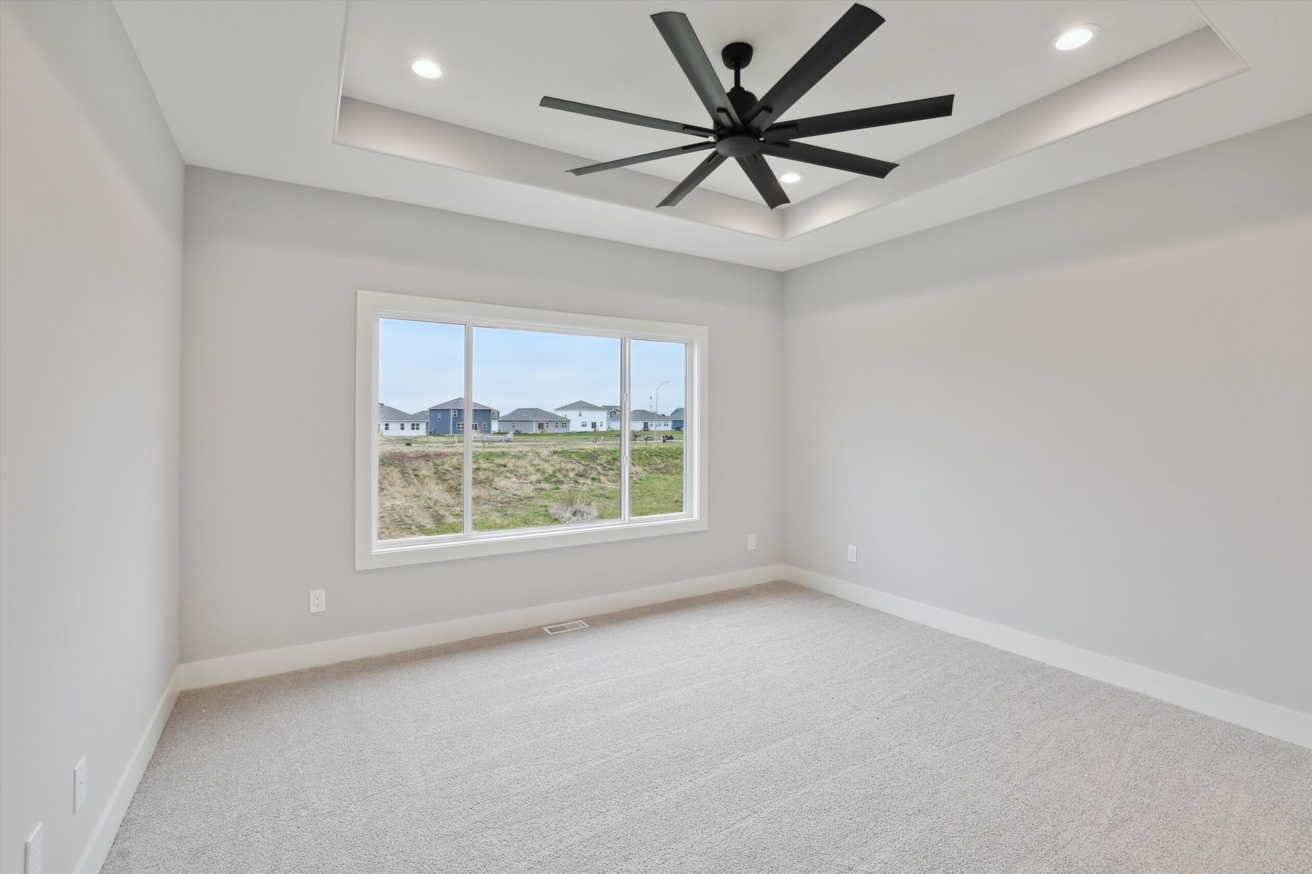 Empty room with beige carpet and light gray walls, featuring a large window with a view of a grassy area and houses. The ceiling has recessed lighting and a modern black ceiling fan. Natural light fills the space.