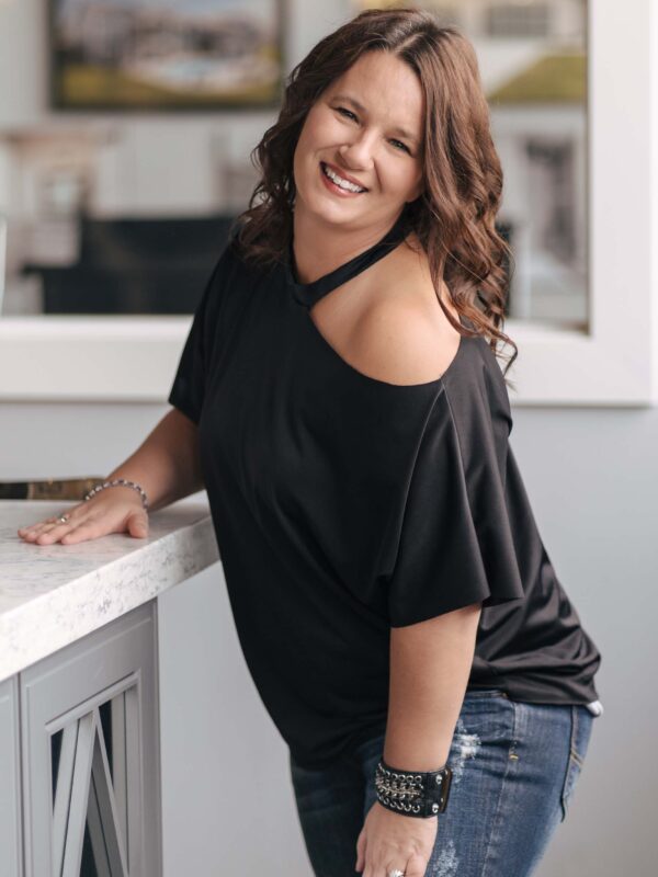 Woman with long brown hair wearing a black off-shoulder blouse and jeans, leaning on a light-colored countertop with a soft smile. The background shows a blurred interior space with neutral tones.