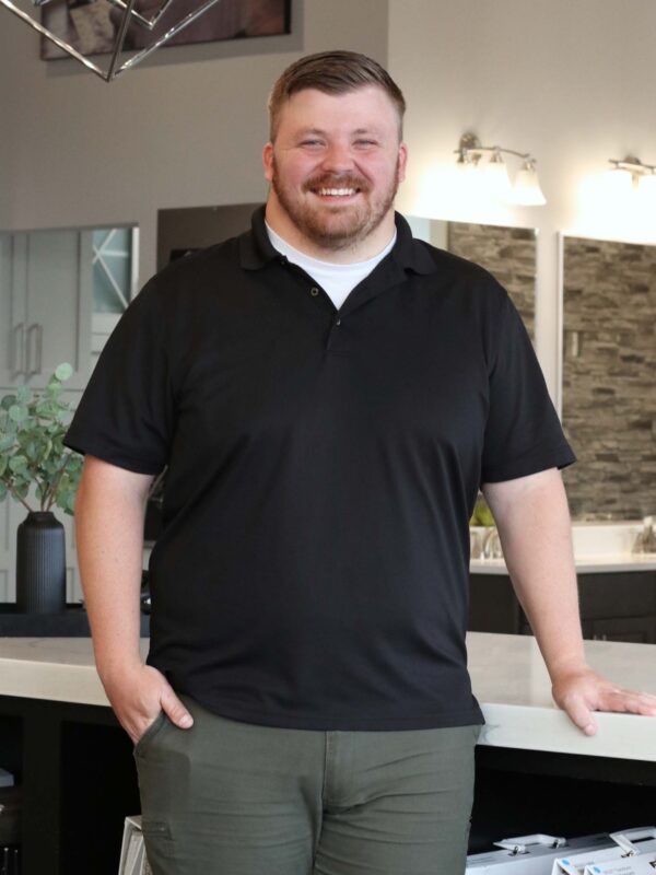 A smiling person stands in a modern showroom with a sleek design. They are wearing a black polo shirt and green pants, with one hand in their pocket. The space features bright lighting and geometric decor elements.