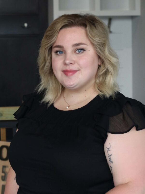 A person with shoulder-length blonde hair and wearing a black top stands indoors. The individual has a neutral expression, with tattoos visible on the arm. A closed cabinet is in the background.