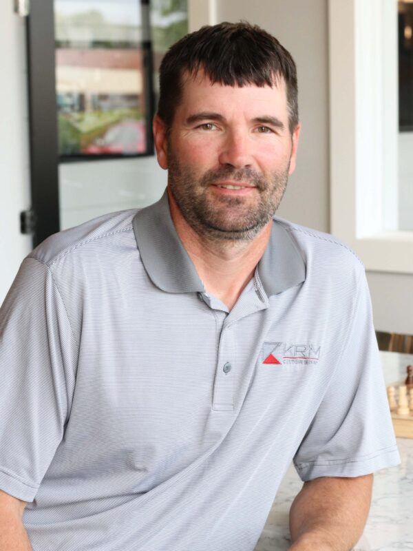 A man with short dark hair and a beard wearing a gray polo shirt with a red and white logo sits at a table. The background includes a window and part of a chair.