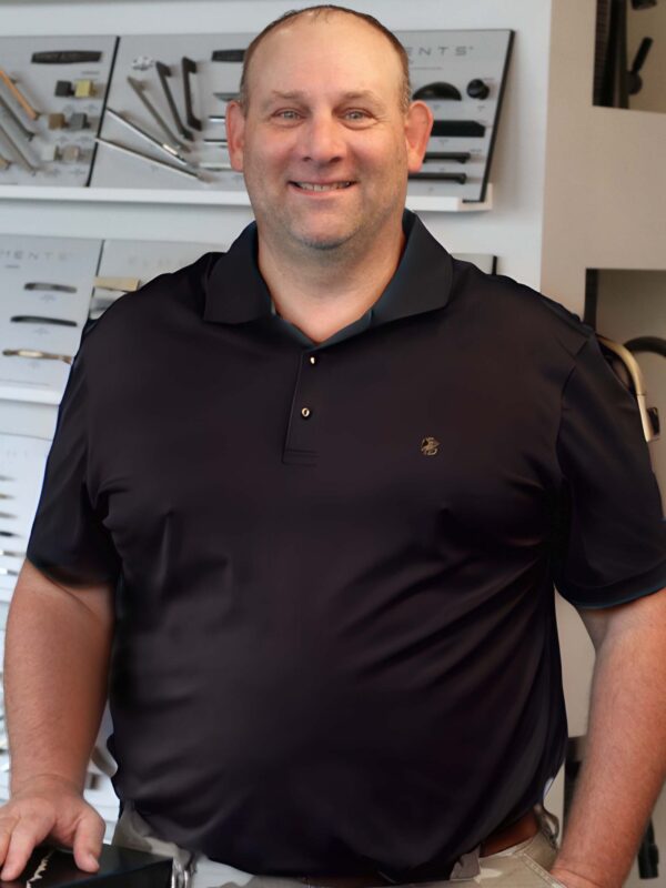 A man in a black polo shirt stands smiling in front of a display wall featuring various metallic hardware pieces.