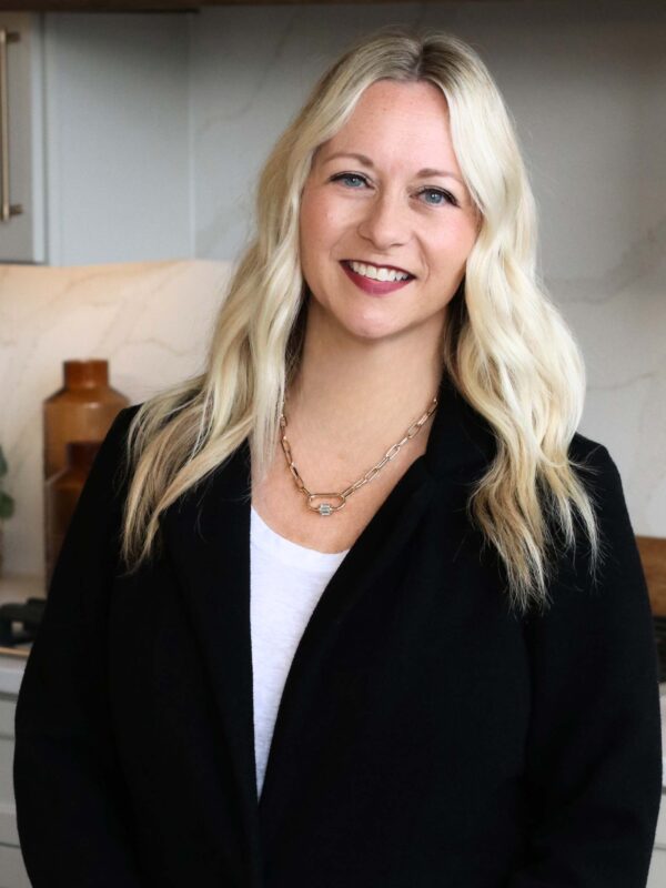 A person with long blonde hair and wearing a black jacket over a white top smiles at the camera. They are standing indoors with a kitchen background featuring light-colored cabinets and decor.