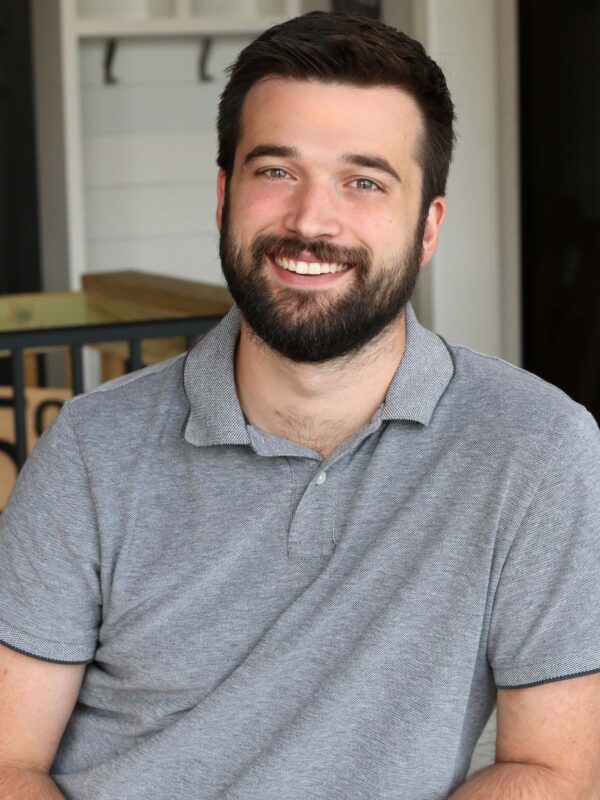 A man with a beard and short hair is sitting and smiling at the camera. He is wearing a gray polo shirt. The background includes a counter and some shelving.