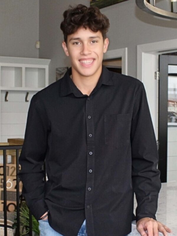 A man with short, curly brown hair is smiling and leaning against a kitchen counter. He is wearing a black button-up shirt and blue jeans. The kitchen has light-colored walls and a wooden shelving unit in the background.