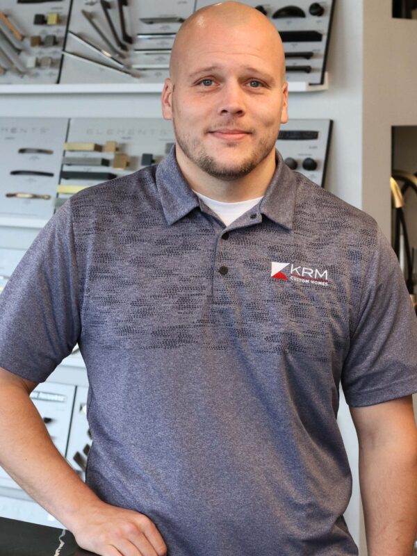 A person in a blue KRM polo shirt stands in a showroom with various product displays on the shelves behind them. The individual has a relaxed expression and is placed slightly to the right in the image.