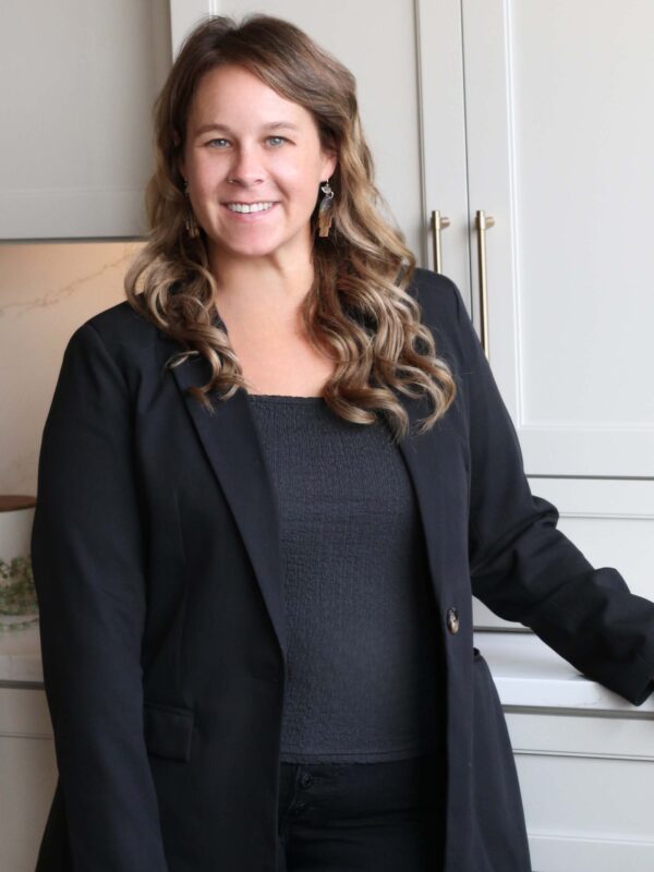 A woman with long, wavy hair is smiling and wearing a black blazer over a dark top. She is standing in front of a light-colored cabinet in a well-lit room.