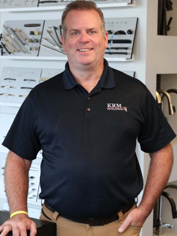 Smiling man in a black KRM polo shirt and khaki pants stands in front of a display wall with various hardware samples.