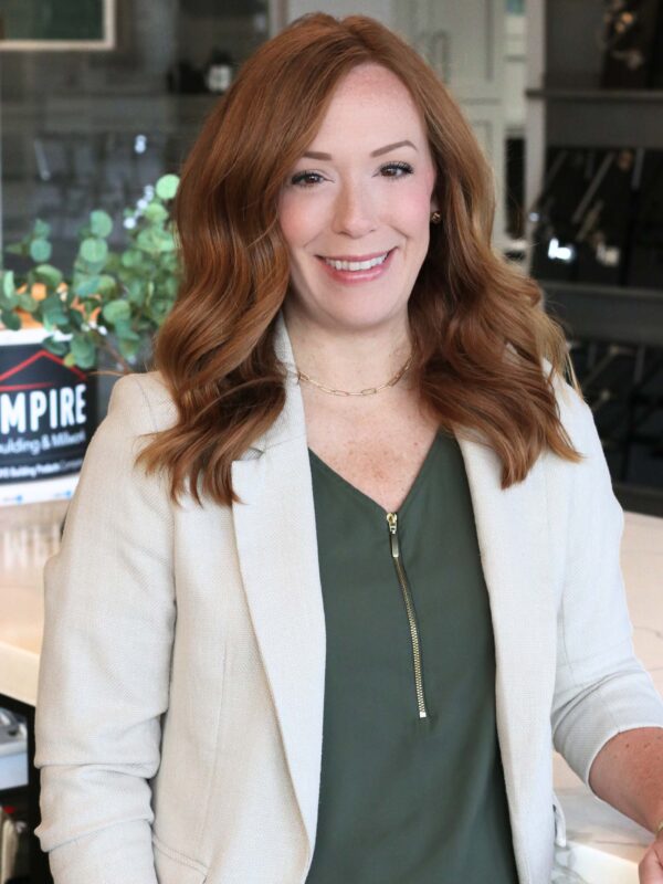 A woman with long, wavy reddish-brown hair is standing indoors, smiling. She wears a white blazer over a green top and is in front of a blurred background with a logo visible.