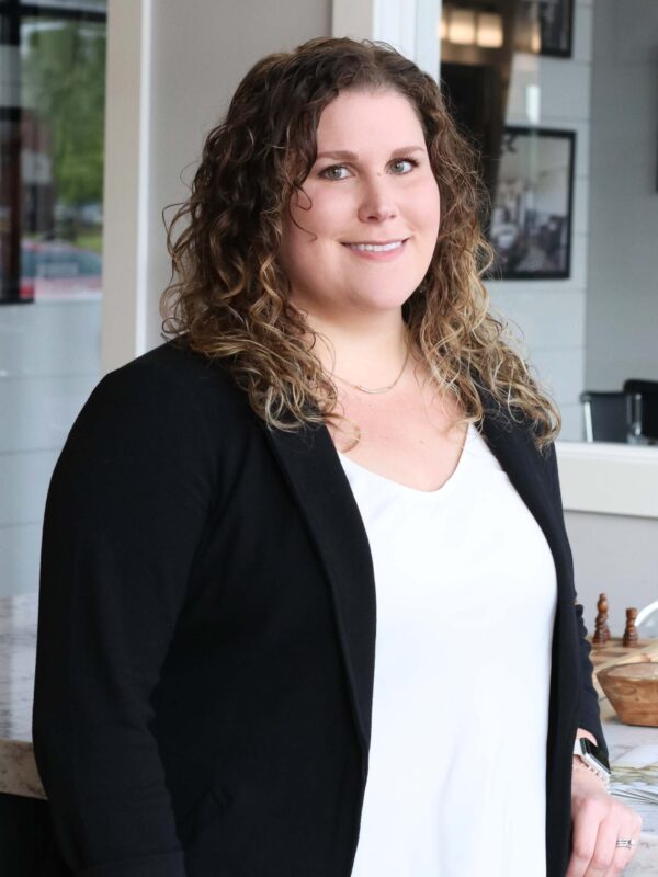 Woman with curly hair wearing a black blazer and white blouse, standing indoors near a counter, smiling at the camera. Background features framed pictures on the wall and a reflective surface.