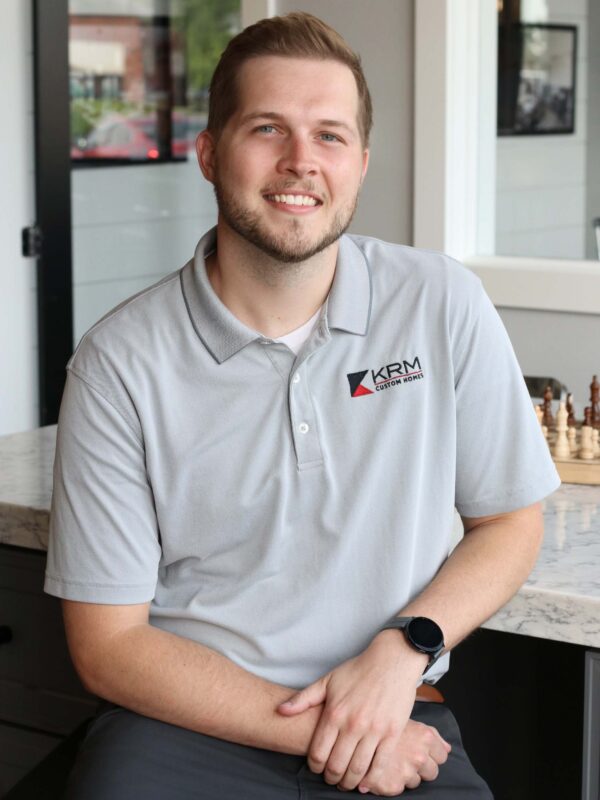 A man with a beard and short hair is smiling and leaning against a marble counter. He is wearing a gray polo shirt with a "KRM Custom Homes" logo. A chessboard is visible in the background.