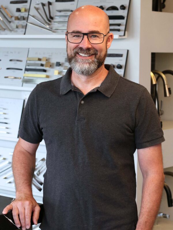 A man with a beard and glasses smiles while standing in front of a display wall showing various handles and hardware. He is wearing a dark polo shirt and has his hand resting on a counter.