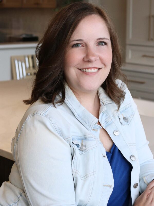 A woman with shoulder-length brown hair smiles warmly while sitting at a table, wearing a light denim jacket over a blue shirt. The background shows a modern kitchen with neutral tones.