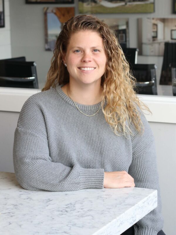 A person with curly, light brown hair smiles while seated at a marble table. They are wearing a gray sweater, with framed pictures on the wall in the background.