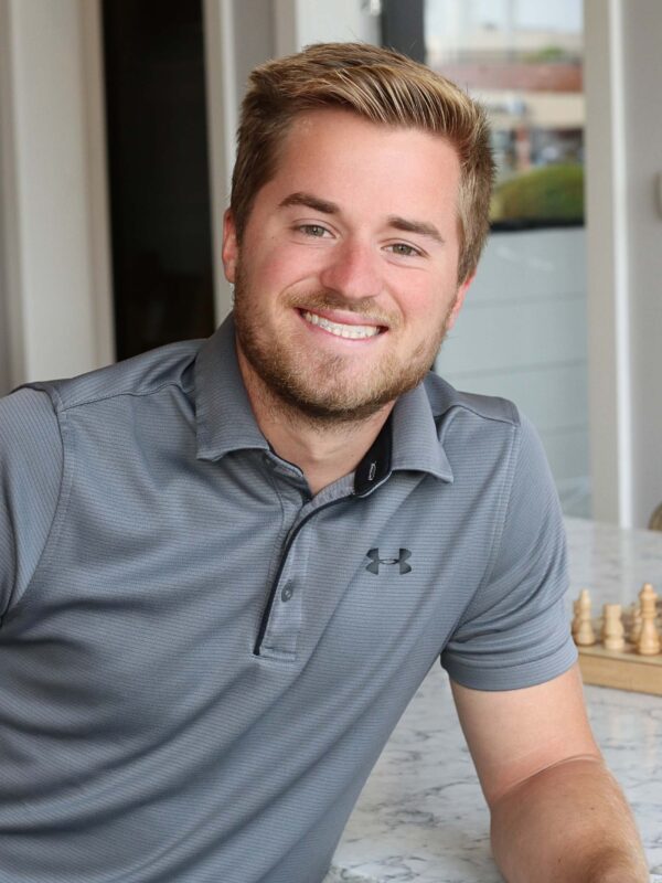 A man with short blond hair and a beard smiles while seated at a table. He's wearing a gray polo shirt. A chessboard is partially visible in front of him, and a doorway is in the background.