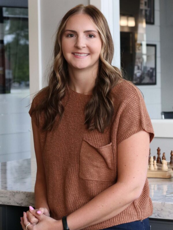 A person with long, wavy hair is sitting on a countertop, smiling. They are wearing a brown knit top and jeans, and a chessboard is partially visible in the background.