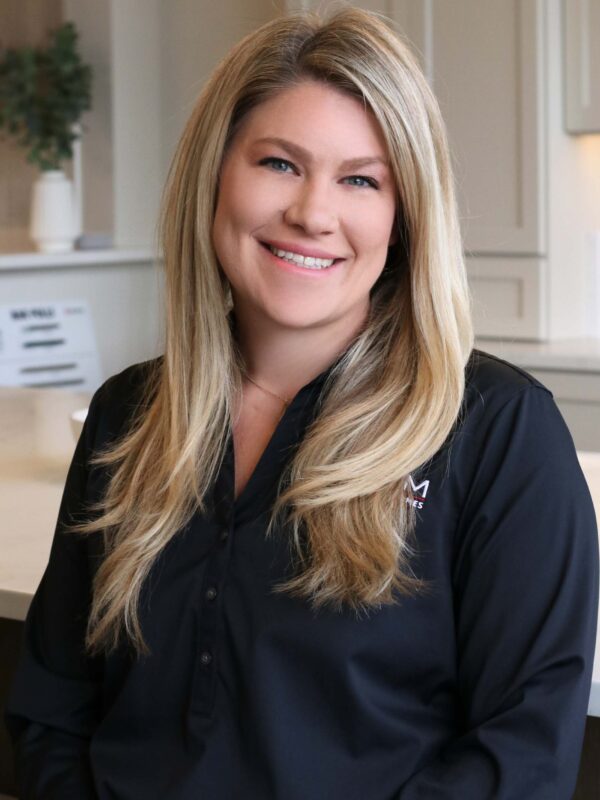 Smiling person with long blonde hair sits in a modern, brightly lit room. They wear a black shirt and are positioned in front of a soft-focus background with greenery and light-colored cabinetry.