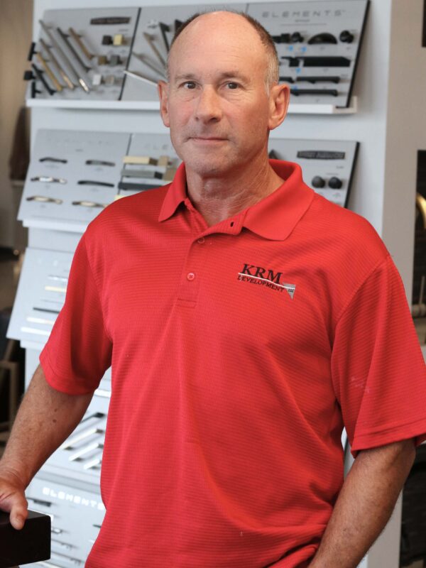 A man wearing a red polo shirt with "KRM Development" logo stands in front of a display featuring various items. He is posing with a neutral expression, and the background is blurry, focusing on the display behind him.