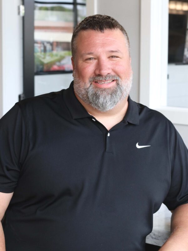 A man with a beard smiles while wearing a black polo shirt with a white logo. He is standing indoors in front of a window and a door.