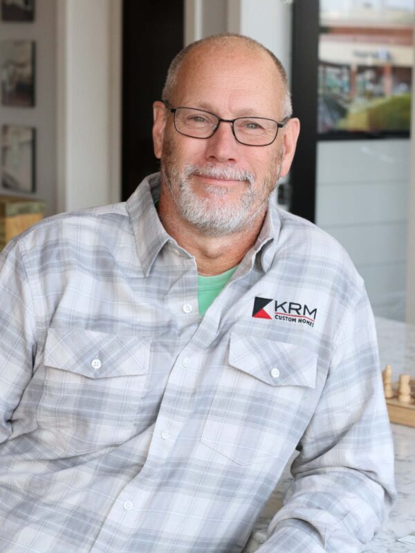 A man with glasses and a beard is smiling. He is wearing a grey plaid shirt with a "KRM Custom Homes" logo. The background shows a modern interior with large windows.