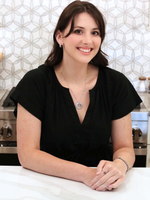 A person with long dark hair smiles warmly, seated at a white counter. They are wearing a black top and a silver necklace. The background features a patterned wall and a silver stove.