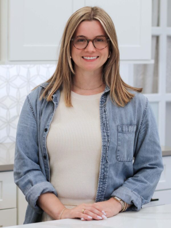 A smiling person with long hair and glasses is standing in a kitchen. They are wearing a denim shirt over a light-colored top. White cabinets and a patterned backsplash are visible in the background.