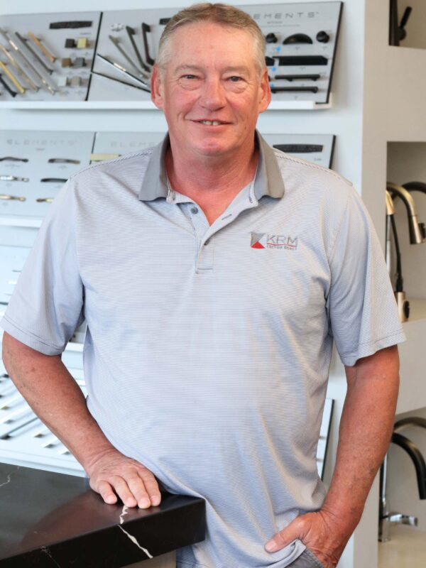 A man in a light gray polo shirt stands with his hand resting on a counter. Behind him are displays of various handles and fixtures. He is smiling and the setting appears to be a showroom or hardware store.