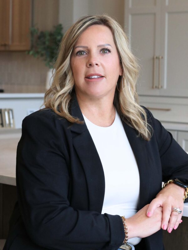 A woman with blonde hair, wearing a black blazer and white shirt, sits in a modern kitchen setting. She has a confident expression with her hands resting on the counter. The background features light wood cabinets and a plant.