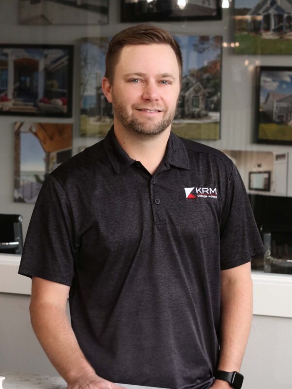 A man with short hair and a beard stands behind a marble counter, wearing a dark KRM Construction Services polo shirt. The background shows framed images of houses on the wall.