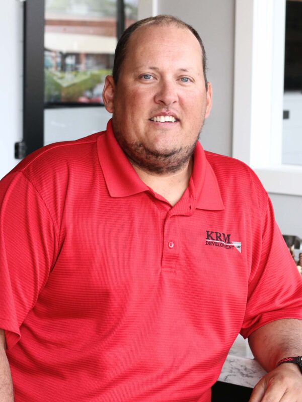 A man wearing a red polo shirt with "KRM Development" logo sits indoors. He has short hair and a light beard, against a background featuring glass windows and a blurred view outside.