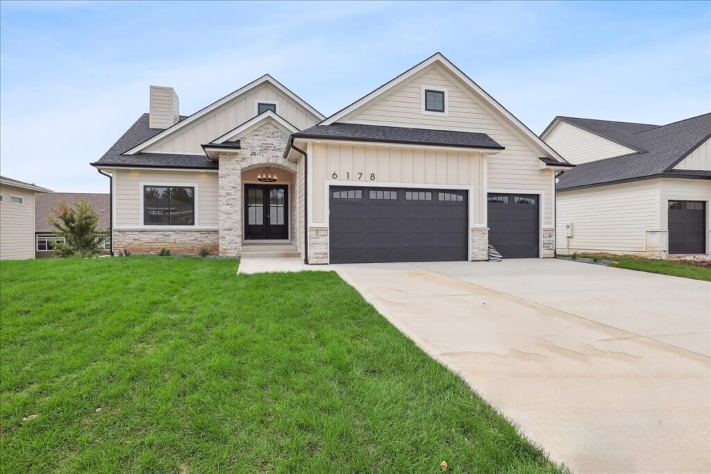 A modern single-story house with light beige siding and stone accents. It features a large driveway leading to two dark garage doors, a small front porch, and a well-maintained green lawn.