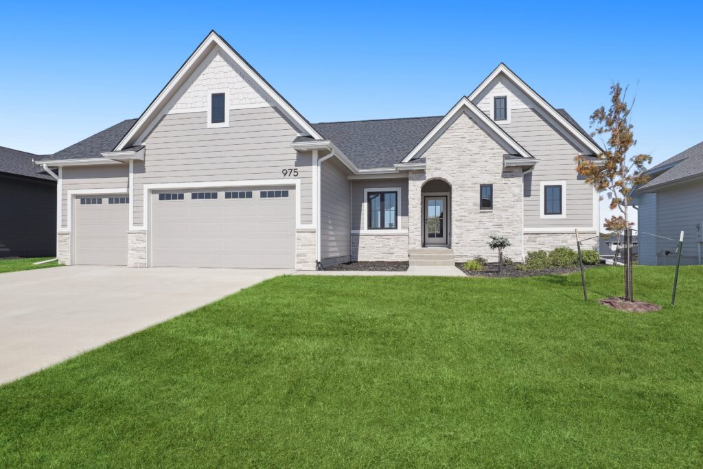 A modern suburban house with light gray siding and stone accents. It features a large driveway leading to a double garage, a small front porch, and a well-manicured lawn under a clear blue sky.