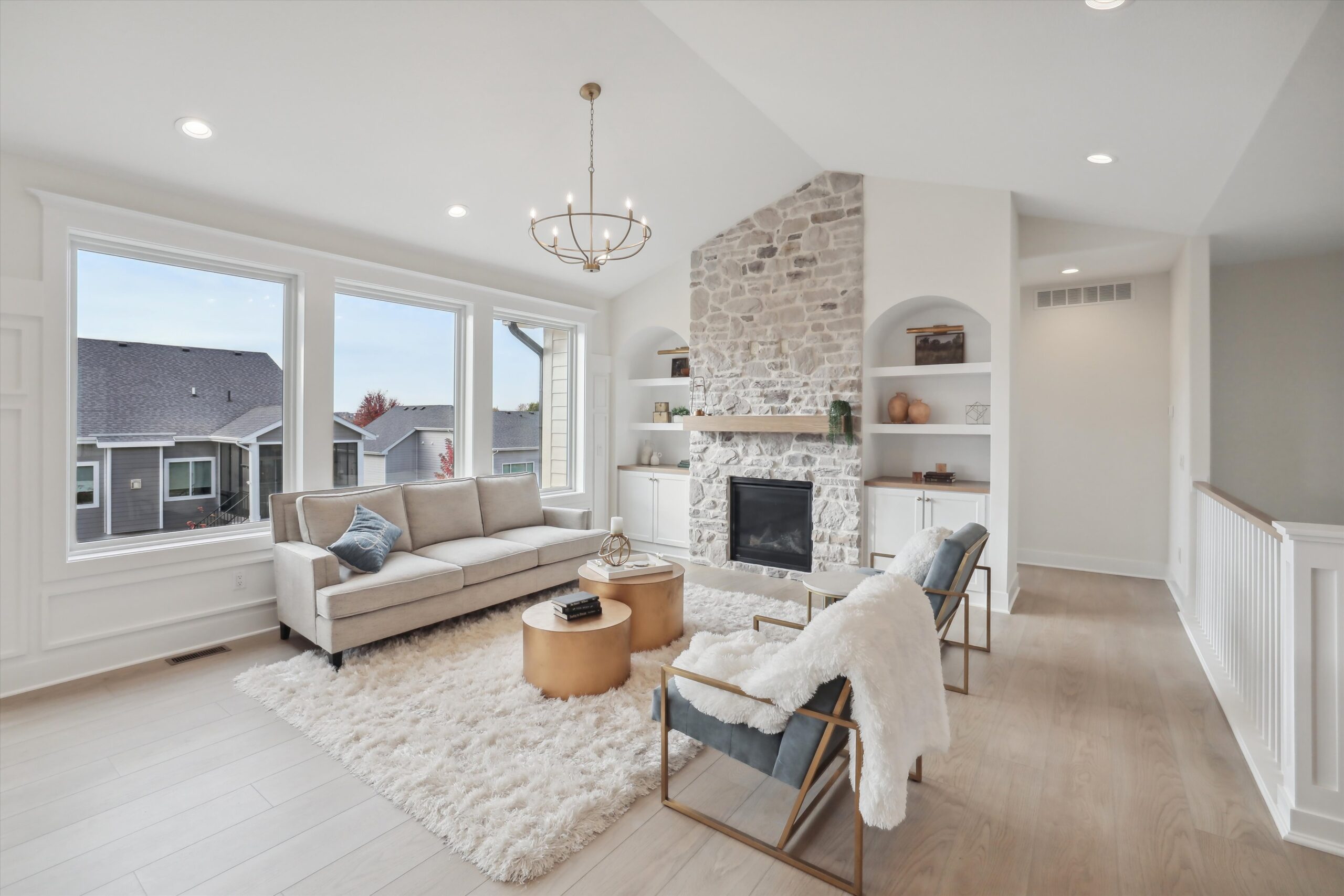 A bright and airy living room with large windows, a stone fireplace, and white walls. There's a beige sectional sofa, two gray armchairs with white blankets, and a round wooden coffee table on a fluffy rug. A chandelier hangs from the ceiling.