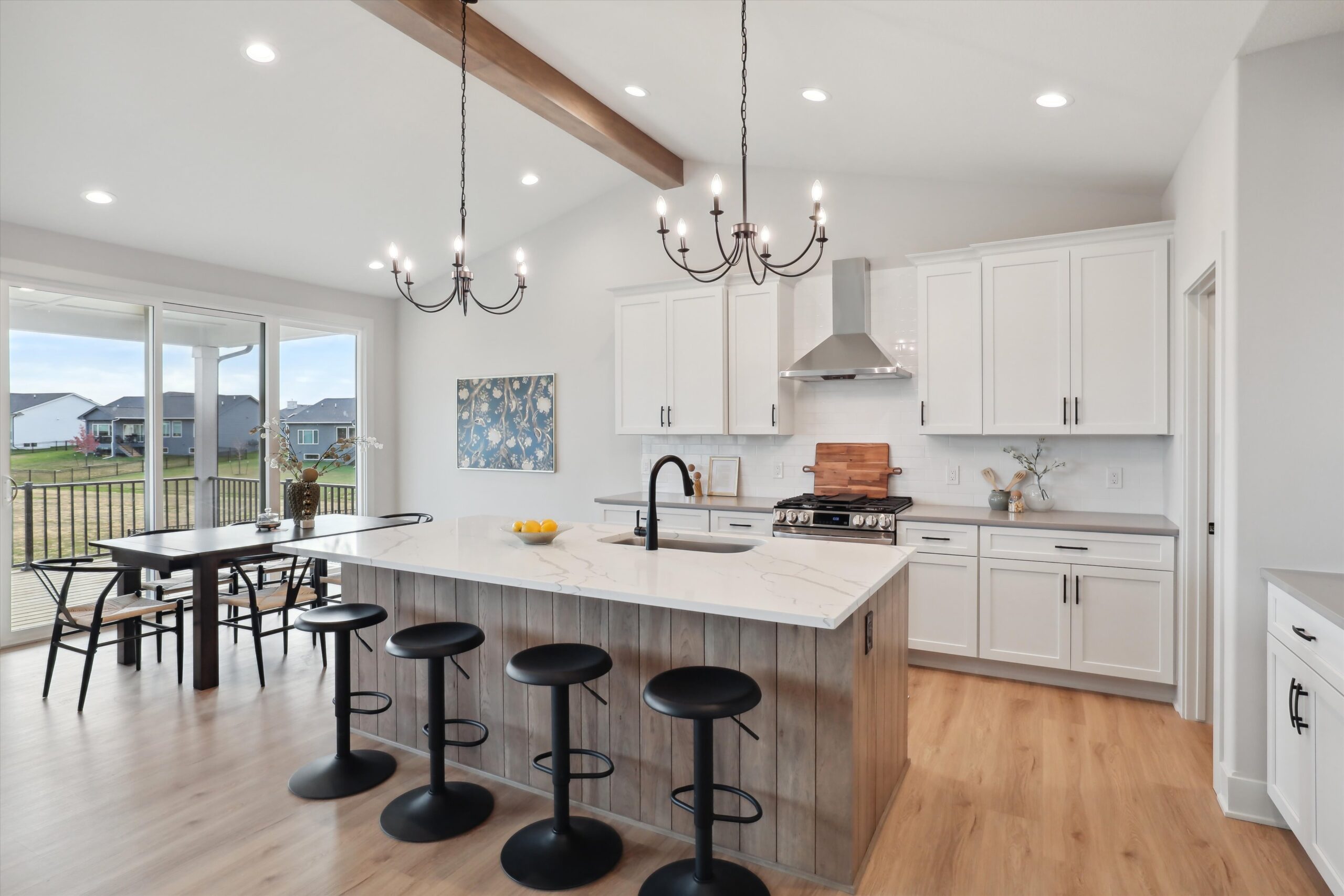 A modern kitchen with white cabinets, a large island with a marble countertop, and four black stools. Two chandeliers hang from the ceiling. There's a dining table near windows that overlook a grassy outdoor view. Light wooden floors complete the space.