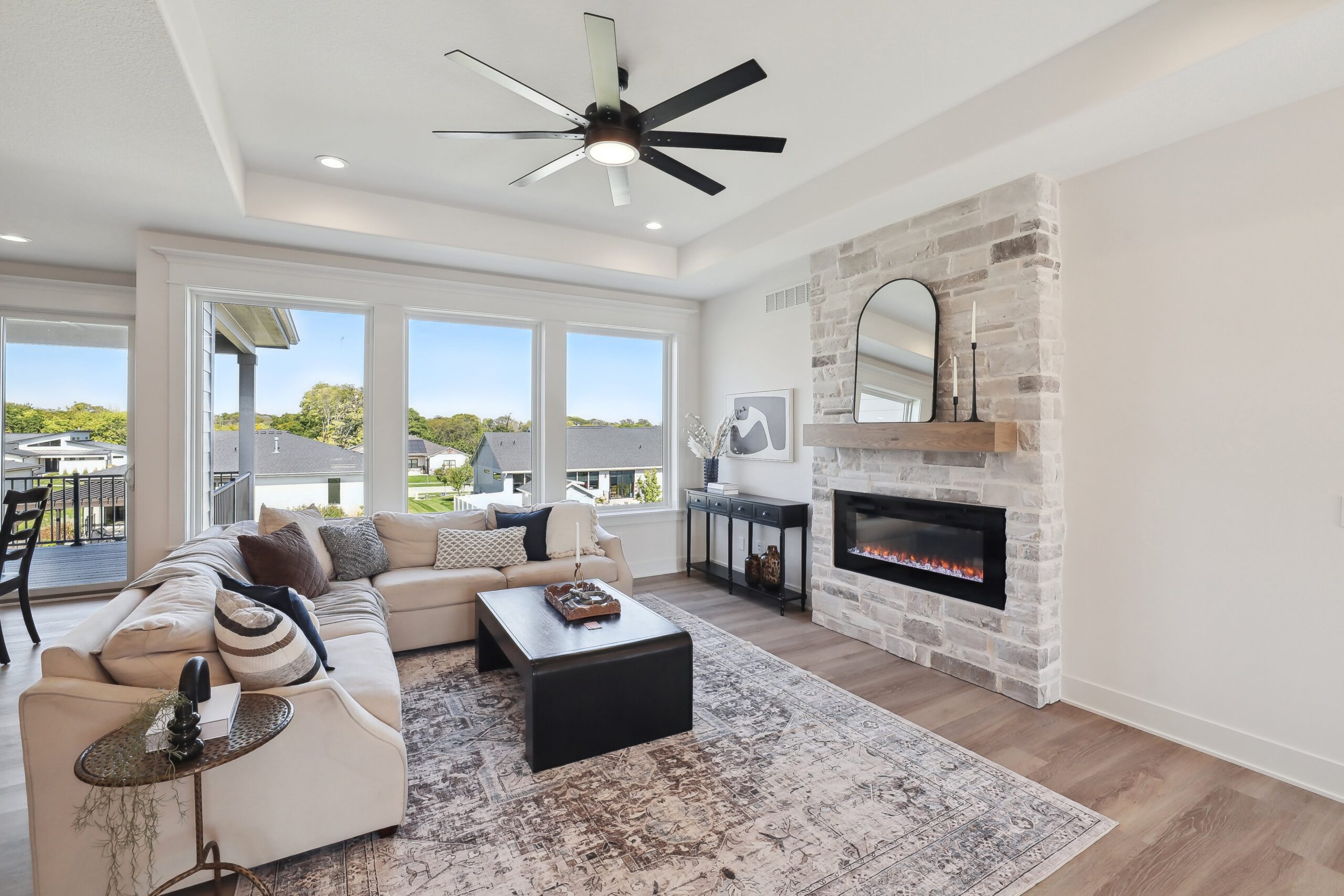 A modern living room with a beige sectional sofa, patterned rug, and a black coffee table. A brick fireplace with a mirror above it is on the right wall. Large windows offer a view of the outdoor deck and greenery.